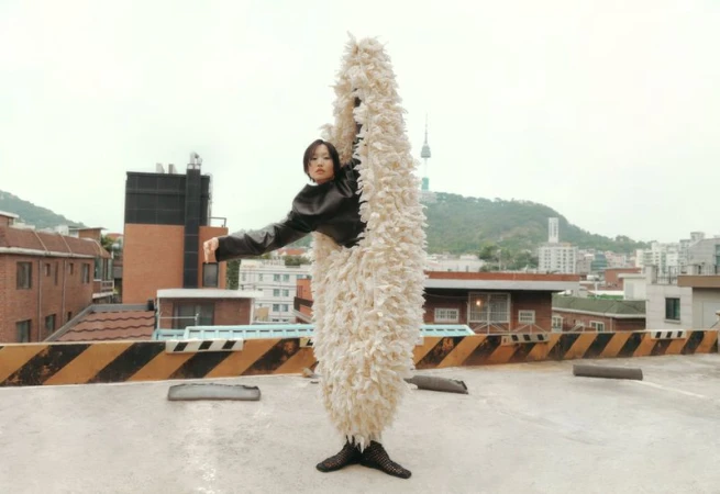 Woman in sculptural costume on a rooftop, Seoul Tower visible in the distance, creating a blend of fashion and urban landscape.