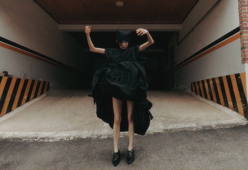 Person wearing an oversized black dress and head covering poses with outstretched arms in a concrete garage or parking structure.