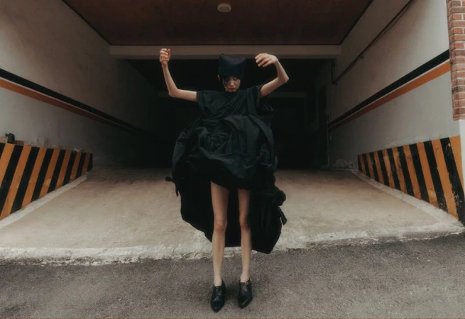 Person wearing an oversized black dress and head covering poses with outstretched arms in a concrete garage or parking structure.