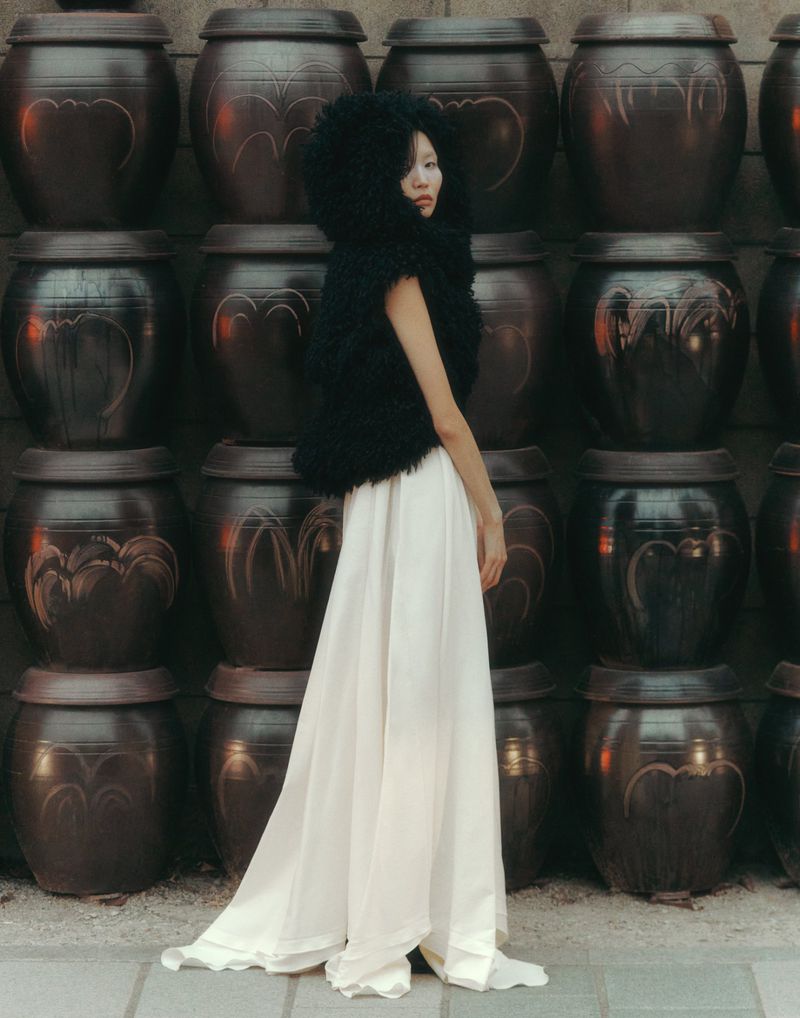 Fashion shot of a woman in a black textured vest and white dress posing in front of a wall of earthenware pots.