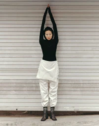 Model standing with arms raised high against a slatted metallic shutter door wearing a black top and white trousers