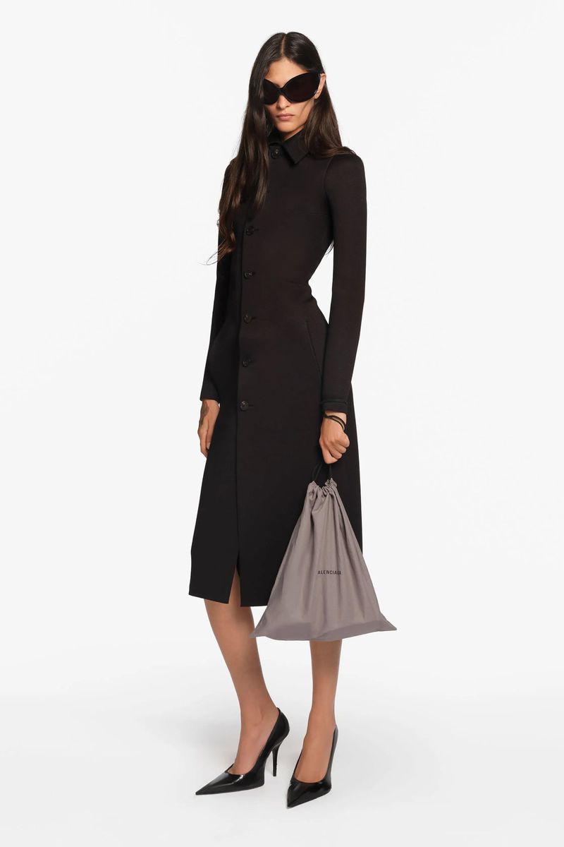 Elegant adult female fashion model in a long-sleeved black midi dress, holding a gray drawstring bag, and posing in a bright photo studio.