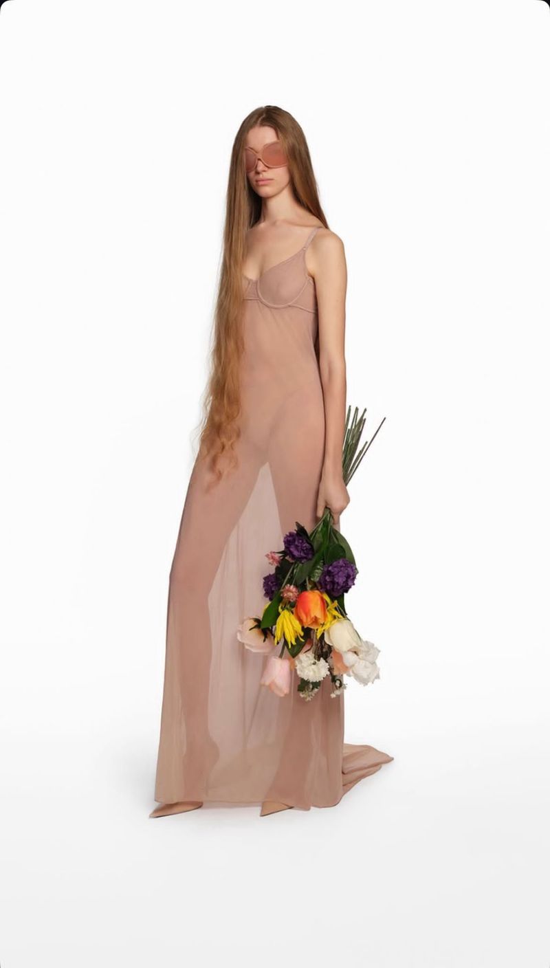 Full-length portrait of an adult female with long hair posing in a sheer dress, holding a bouquet of flowers on a white backdrop.