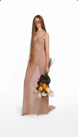 Full-length portrait of an adult female with long hair posing in a sheer dress, holding a bouquet of flowers on a white backdrop.