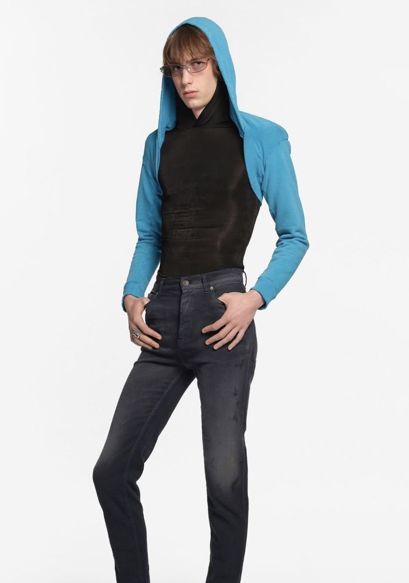 Adult male poses in a blue hoodie and fitted black top with dark jeans against a white backdrop in a fashion studio shot.