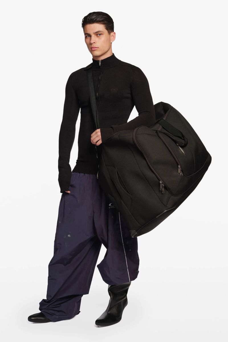 An adult male fashion model stands indoors in a photo studio, wearing a black top and baggy blue pants, carrying a large black bag.