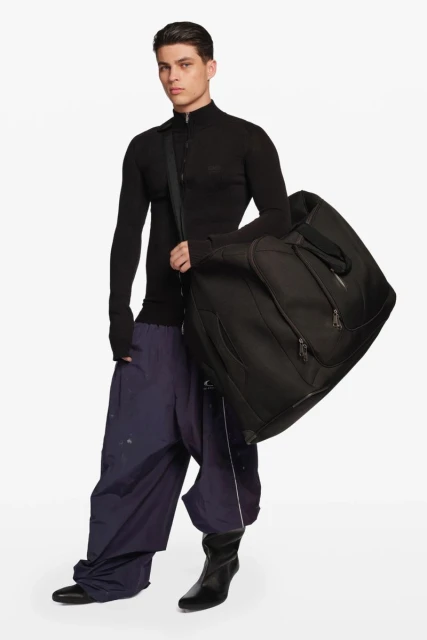An adult male fashion model stands indoors in a photo studio, wearing a black top and baggy blue pants, carrying a large black bag.
