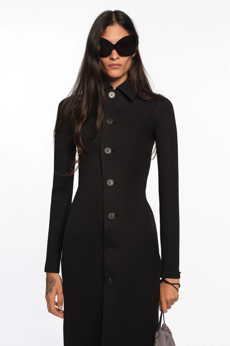 Woman wearing a black dress and butterfly sunglasses poses in a bright studio. The garment is long-sleeved with buttons.