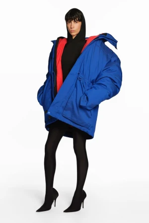 A woman models a blue oversized puffer jacket with a red interior, black hoodie, leggings, and high heels against a white backdrop.
