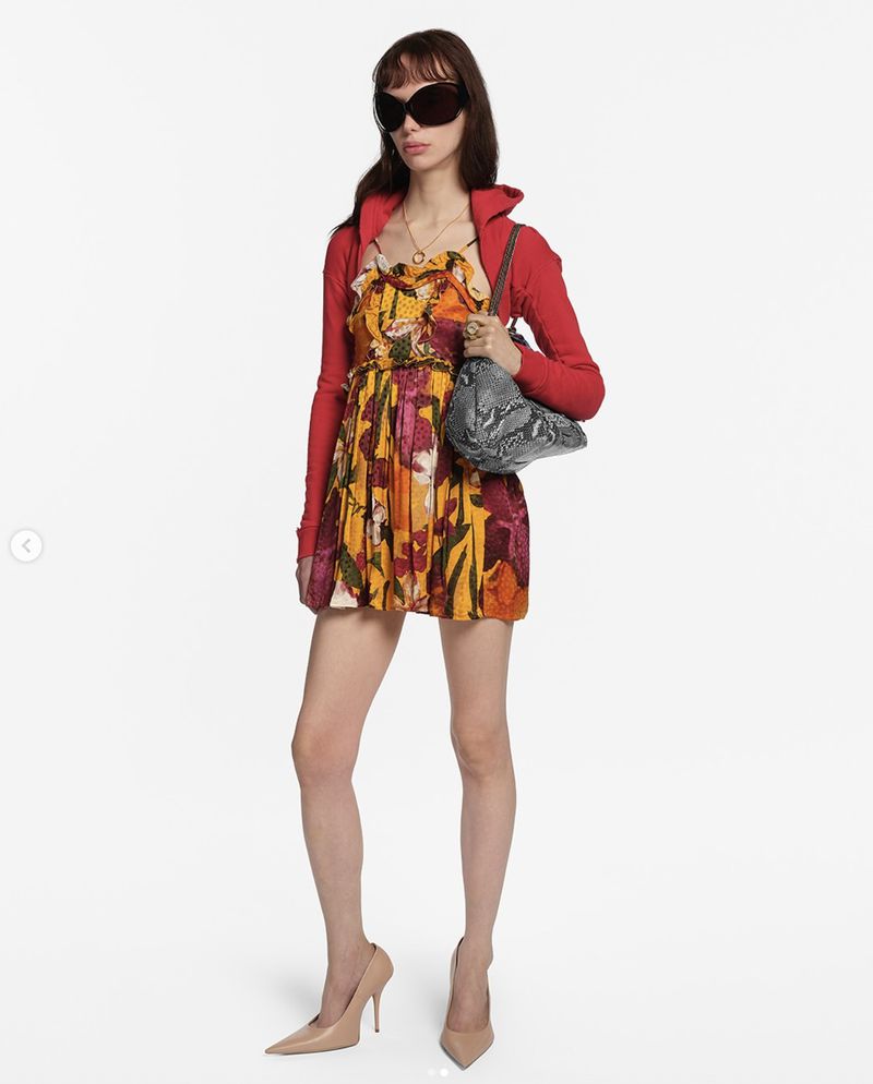 A woman in a floral dress, red jacket, heels, and sunglasses poses in a full shot against a white background, carrying a patterned handbag.