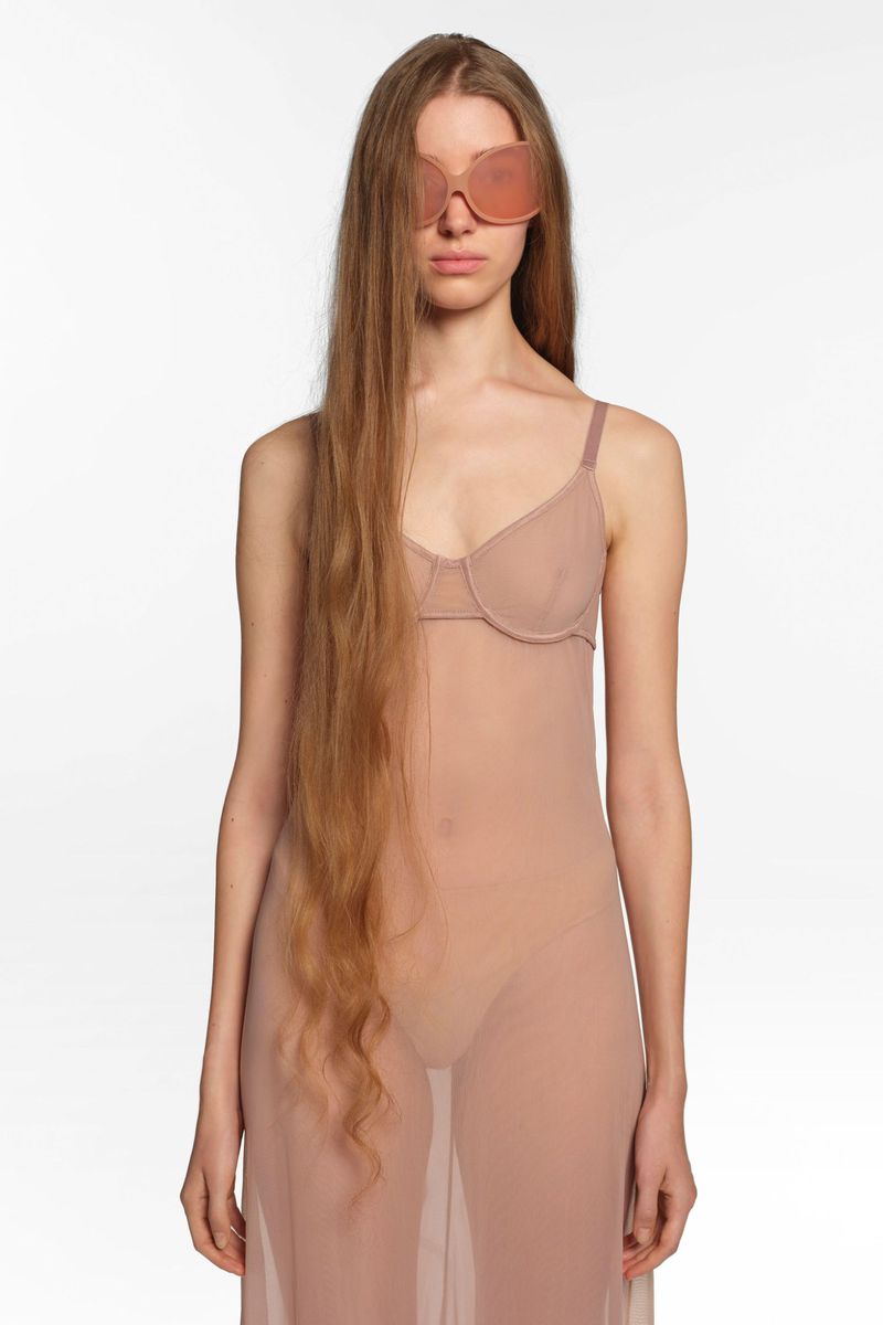 A woman poses in a sheer dress with oversized sunglasses on a white background, showcasing fashion and beauty in a studio setting.