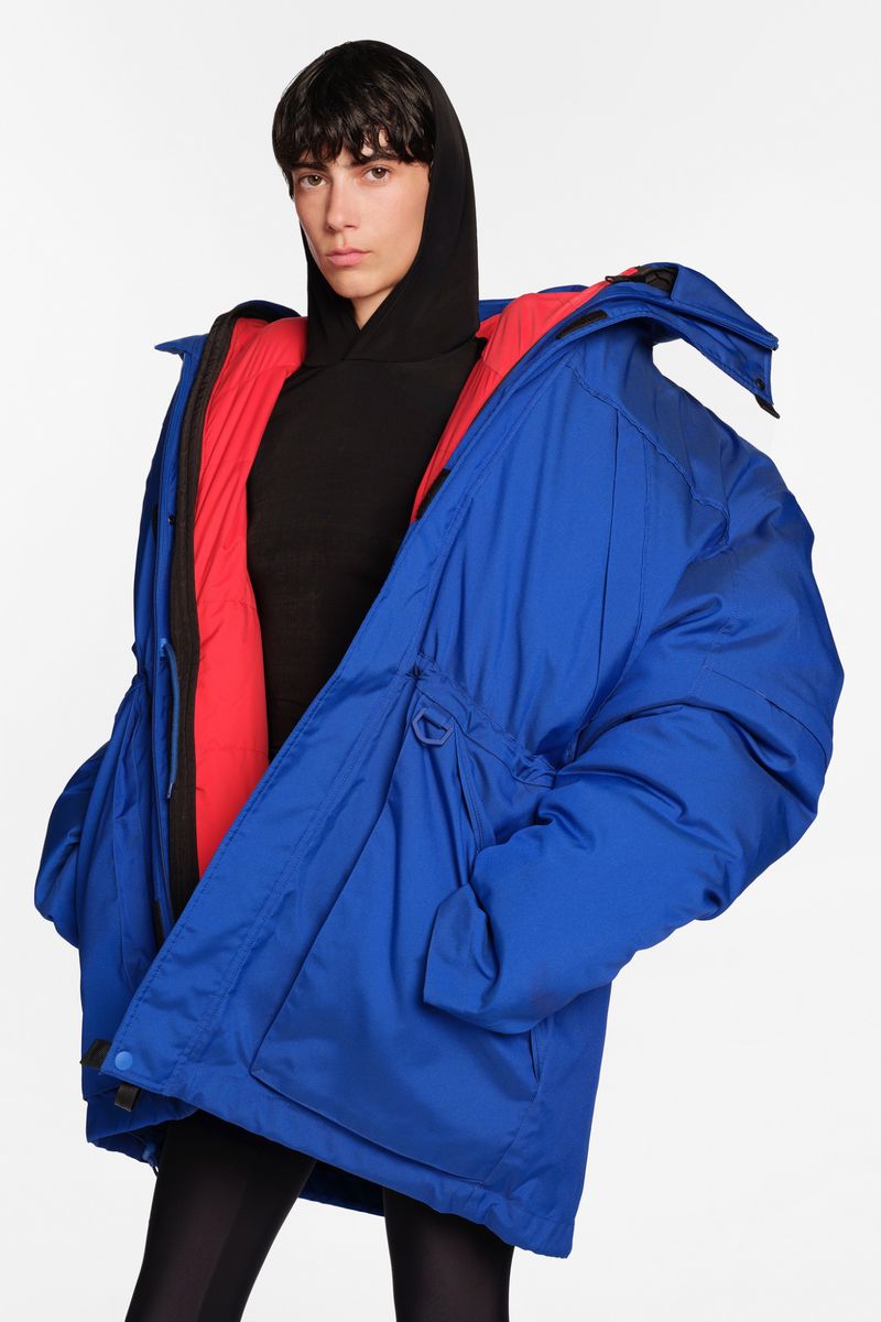 Fashion shot of a model wearing an oversized blue puffer jacket with a red interior and a black hoodie, posing against a white backdrop.