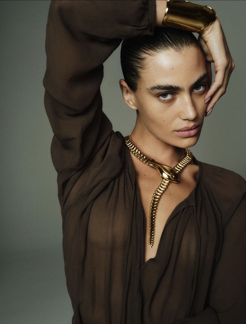 Close-up of a young adult woman with a snake necklace and gold cuff. Wearing a brown chiffon top and posing in front of a grey background.