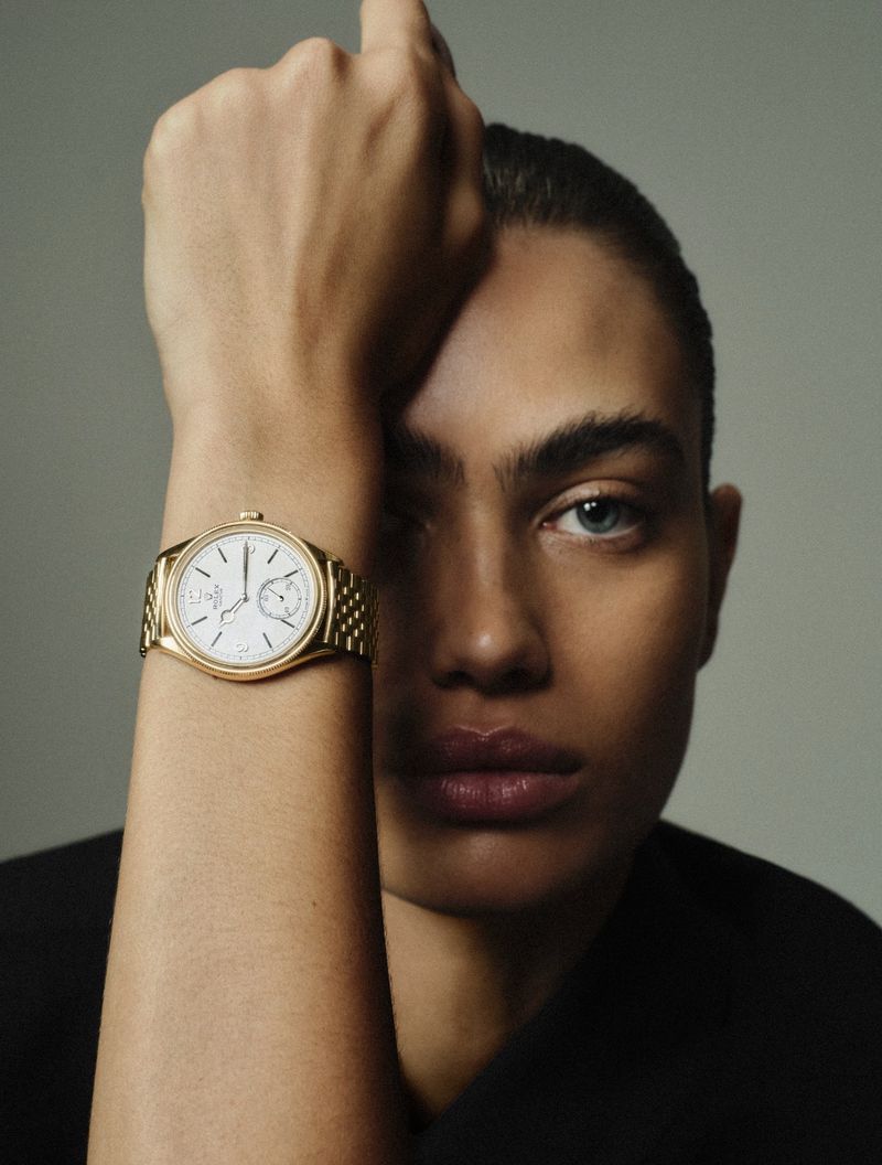 Close-up of an adult with light skin, showcasing a luxury gold wristwatch with a white dial against a neutral grey backdrop.
