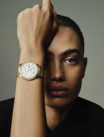 Close-up of an adult with light skin, showcasing a luxury gold wristwatch with a white dial against a neutral grey backdrop.