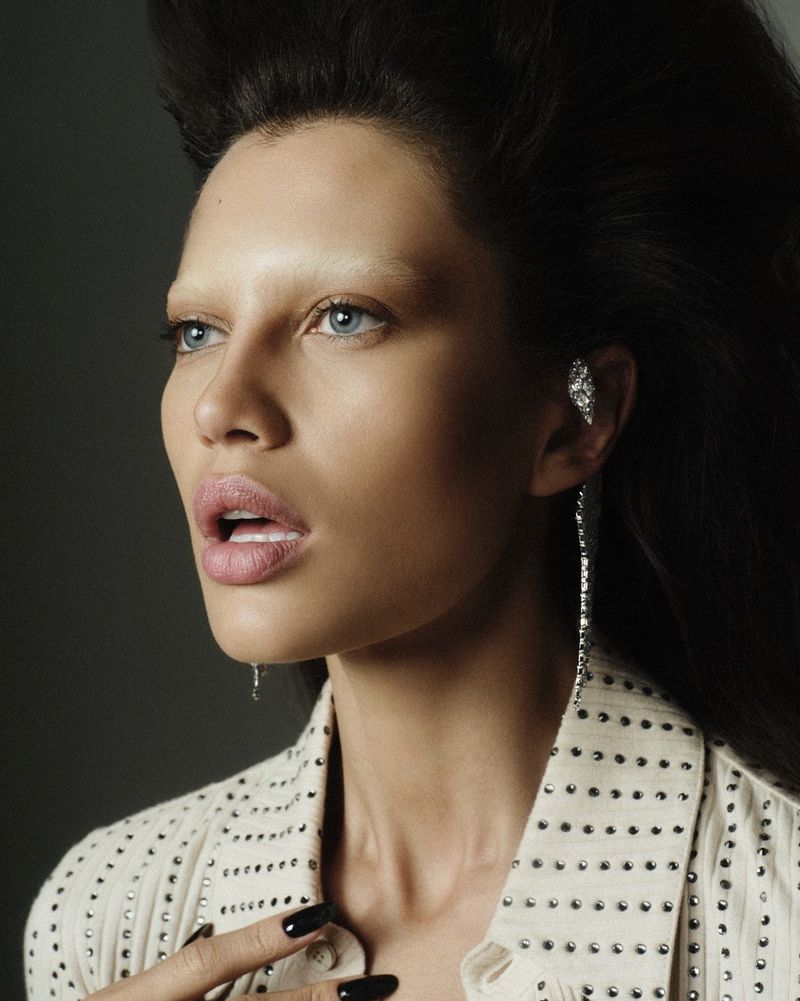 Close-up portrait of an adult female model with dark hair, light skin, light brows, wearing a studded jacket and diamond ear cuff.