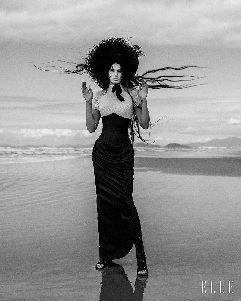 Editorial fashion shot of a woman on a beach in a black dress and large dark hat against a cloudy sky and the ELLE magazine logo.