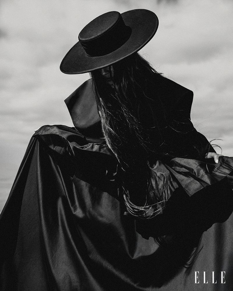A single female model in a wide-brimmed hat and dark dress posing dramatically against a cloudy sky for a black-and-white fashion editorial.