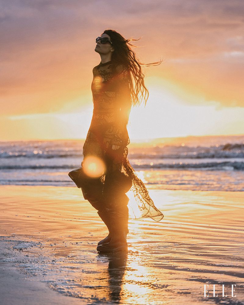 Woman in sheer dress and sunglasses walks on beach at sunset with golden light and gentle waves, reflecting on the wet sand.
