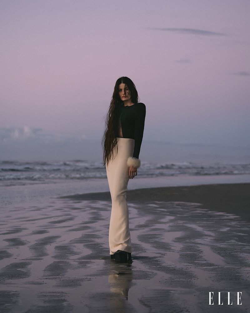A young woman with long dark hair stands on a wet sandy beach in a long white skirt and dark top at dusk, with ELLE logo.