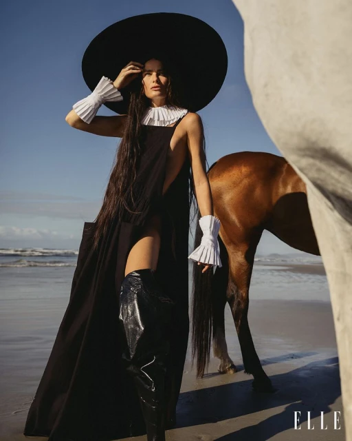 Woman in wide-brimmed hat poses with a horse on sandy beach with water and blue sky in the background, fashion photography.