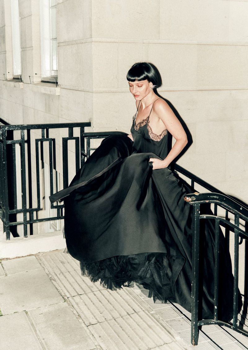Adult female descends stairs in a long, dark gown with lace trim and bob haircut, captured in an indoor setting with soft lighting.