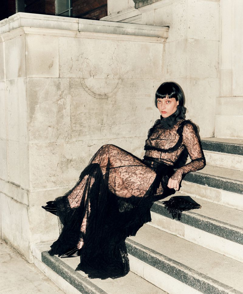 Woman in a black lace dress sits on stone steps, gazing at the camera, with glossy dark hair and a black accessory in her hand.