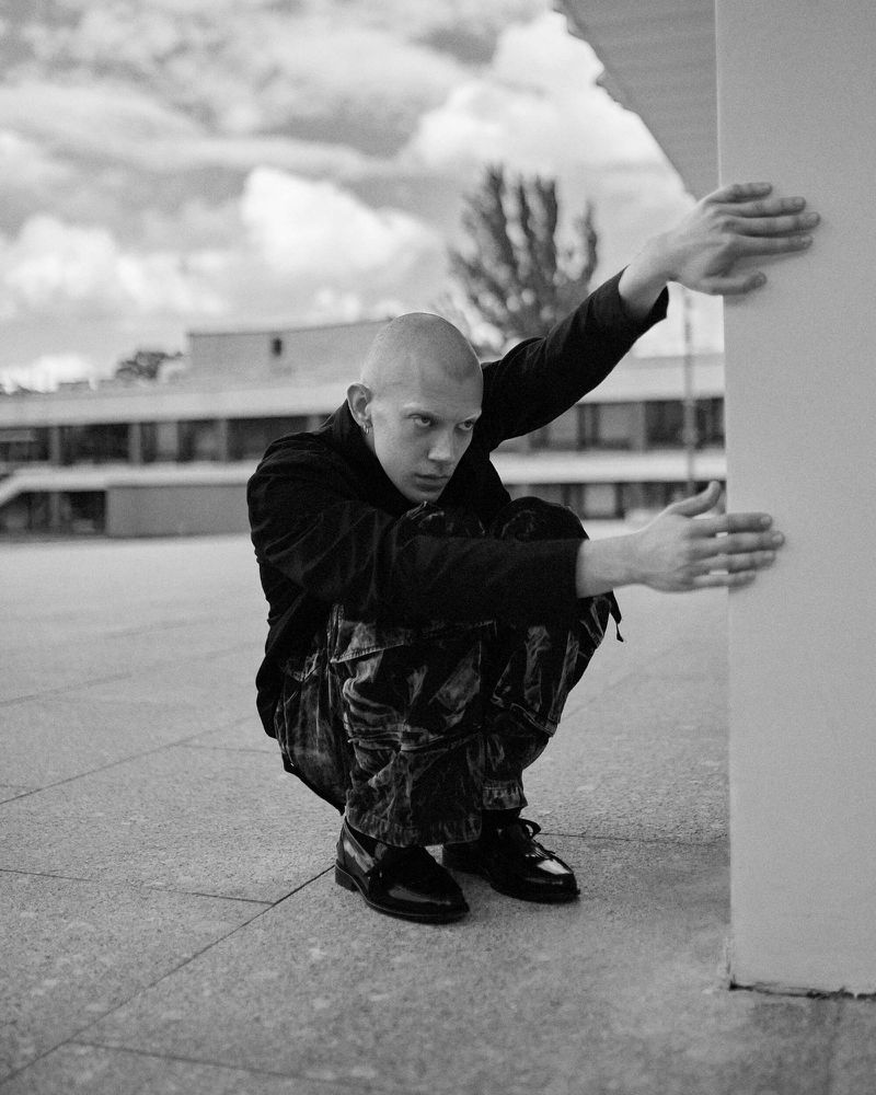 Monochromatic portrait of a bald adult crouching in a courtyard, wearing dark clothing, and posing next to a white pillar in daylight.
