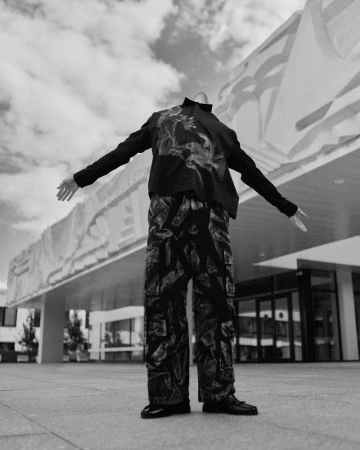 Monochrome low-angle editorial photograph of a headless figure wearing patterned cargo trousers and a black long-sleeve shirt outdoors.