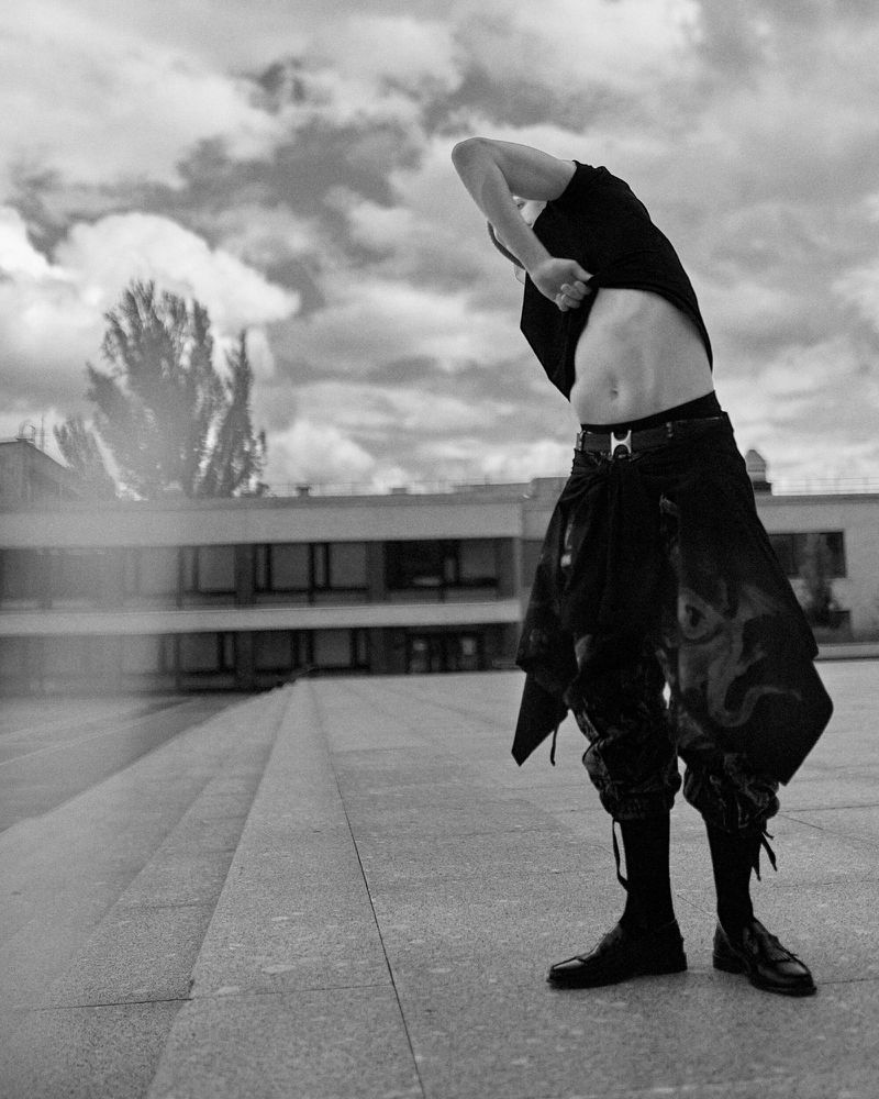 Monochrome image of a shirtless man in baggy pants standing on a rooftop, slightly bending back with his hand up. Cloudy sky background.