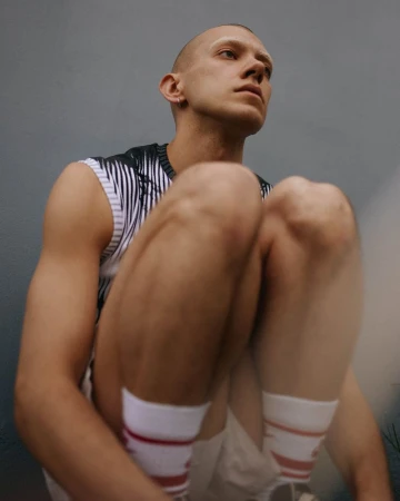 Portrait of a fair-skinned young adult male with a shaved head and striped tank top sitting with legs bent, wearing socks with red stripes.