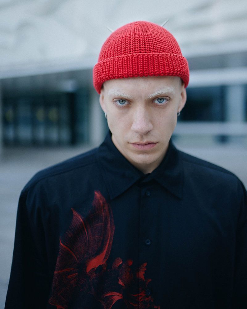 Portrait of an adult male with light skin and a red beanie, wearing a black button-down shirt with a red floral design, posing outdoors.
