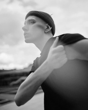 A close-up black and white portrait of a person in a black beanie and black t-shirt looking away with a serious expression.