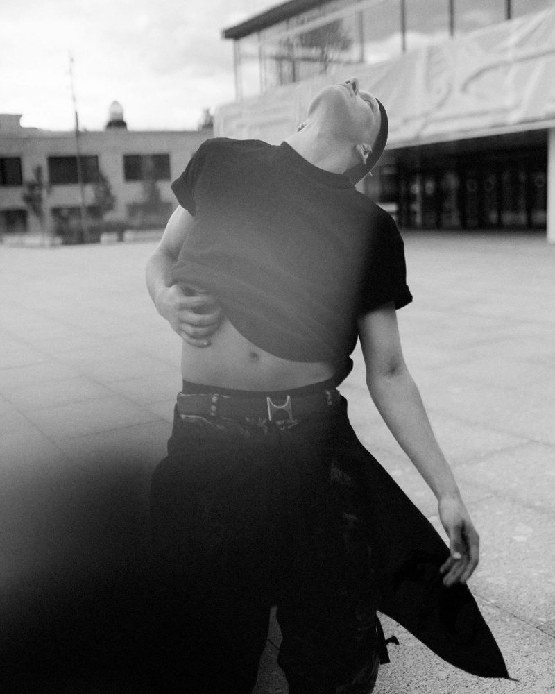 Monochrome shot of a slender adult male in an outdoor urban setting, tilting his head back and lifting his shirt in black pants.