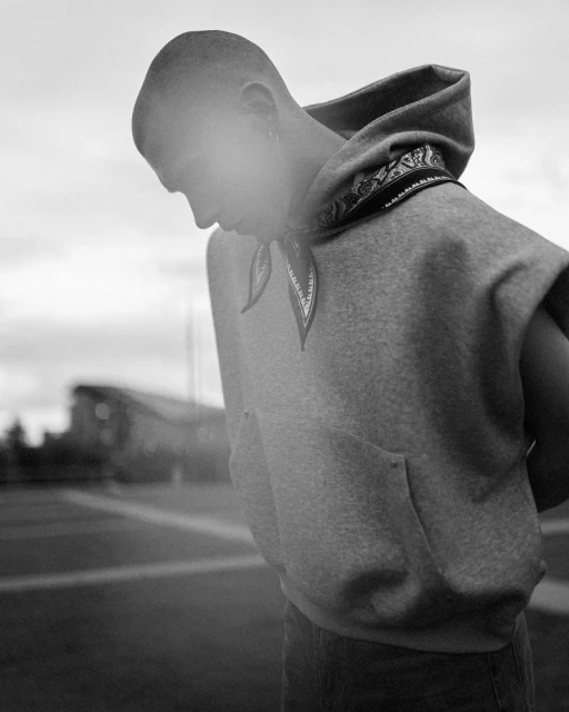 Monochrome portrait of a young adult male in a sleeveless hoodie with a bandana detail outdoors against a blurred background.
