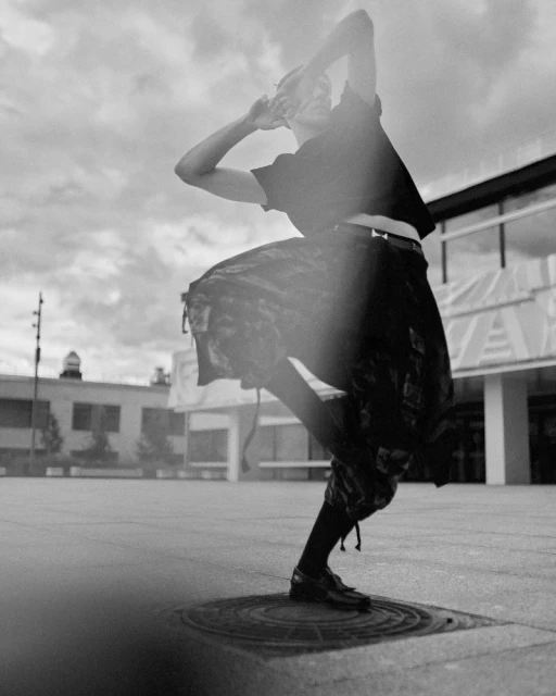 Black and white shot of a person in a dark top and patterned skirt striking a dynamic pose on a city square with cloudy sky above.