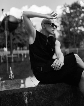 Monochrome portrait of a seated bald man in black clothing shielding his eyes outdoors