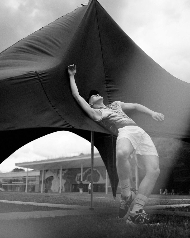 Monochrome portrait of a man in shorts leaning against a black tent on a grassy field with a building in the background.