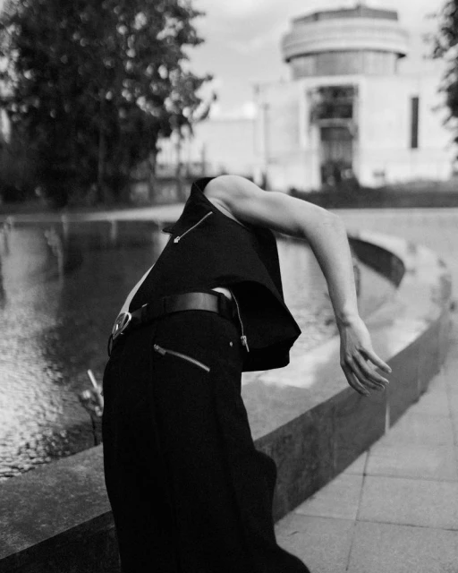 Black and white photograph of a human figure in dark clothing bending backwards outdoors, with an out-of-focus building in the background.