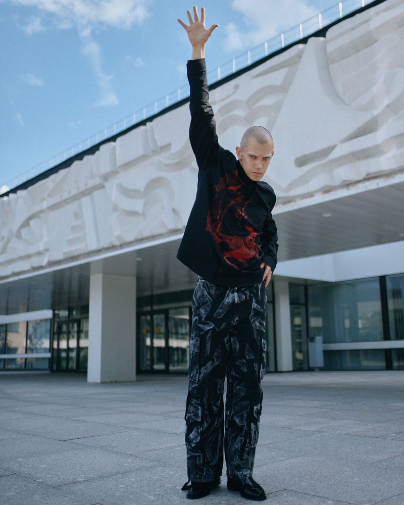 Young adult poses in black patterned clothing outside a modern building under a bright blue sky, raising his hand overhead.