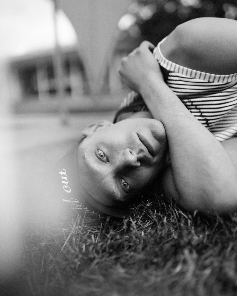 Black and white portrait of a person with blue eyes lying in grass wearing a striped shirt, shot at eye-level and shallow depth of field.