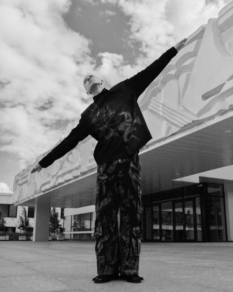 A person stands with arms spread wide in front of a modern concrete building, wearing a black shirt and patterned cargo pants.