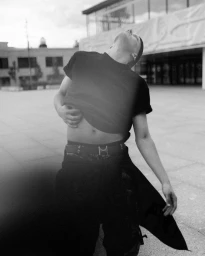 Monochrome editorial portrait of a person in a black t-shirt pulling up their hem outdoors in front of a building.