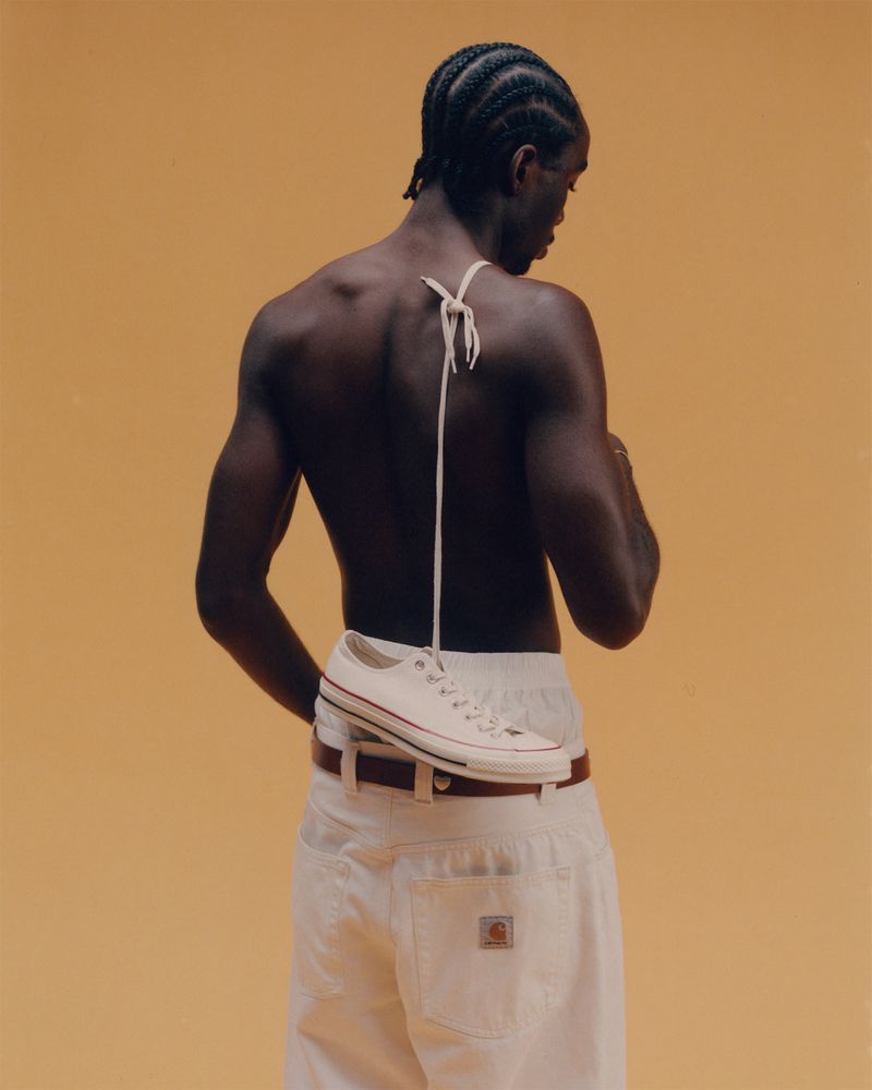 Back view of an adult male wearing white jeans, a belt, and holding a white sneaker on his back against a light-orange background.