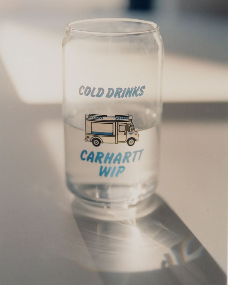 A glass with a Carhartt logo and cold drinks text, partially filled with a clear liquid, casting a shadow on a white surface.