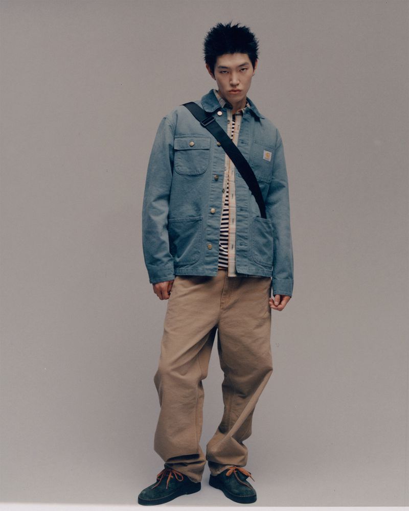 Full-length studio shot of a young adult male fashion model wearing a blue Carhartt jacket and khaki pants against a neutral backdrop.