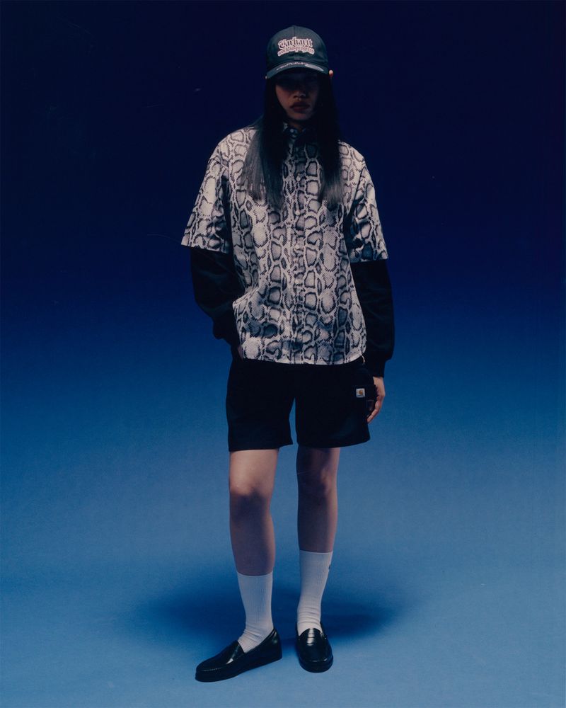 A full shot of a person in a snakeskin-print shirt, black shorts, white socks, and black loafers posing against a graduated blue backdrop.