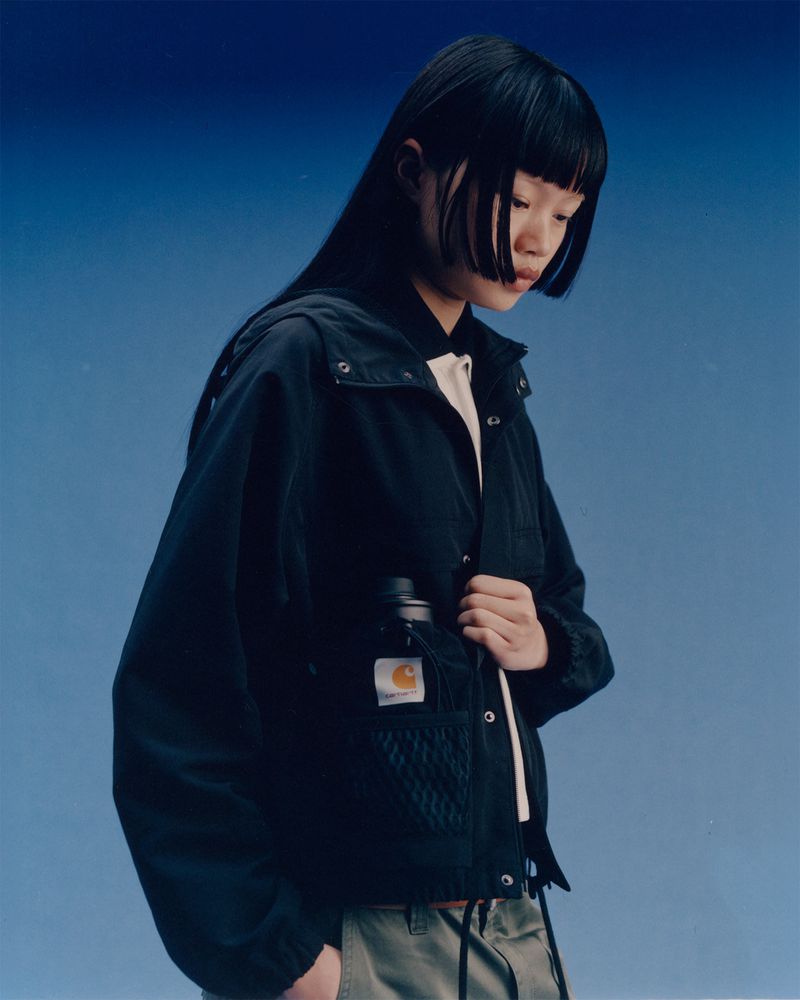 Asian woman in Carhartt jacket with water bottle against blue gradient background. Fashionable jacket and neutral color palette.