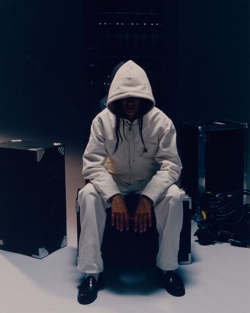 Person in white hooded jacket and pants sitting on a black road case indoors in a dim photo studio with black leather shoes.