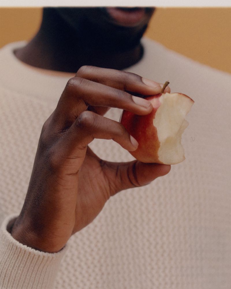 Close-up of a dark-skinned person holding a bitten red apple against a yellow background, wearing a cream-colored sweater.